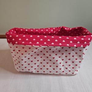 May include: A rectangular fabric basket with a red and white heart pattern. The basket's interior and trim are a solid red with white hearts. The exterior is white with small red hearts. The basket is made of fabric and is likely used for storage.