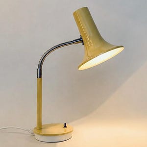 Puede incluir: Una lámpara de escritorio amarilla con una pantalla cónica acampanada, un brazo flexible de acabado cromado y un poste cilíndrico amarillo unido a una base redonda amarilla. Un pequeño interruptor de palanca negro se sitúa en la base.