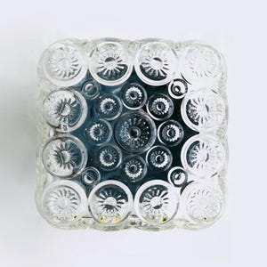 Può includere: Apparecchio di illuminazione quadrato in vetro trasparente con un motivo di disegni circolari a forma di fiore. L'apparecchio ha un interno scuro ed è montato su uno sfondo bianco. Il vetro ha un aspetto strutturato.