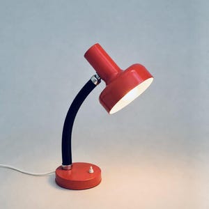 Puede incluir: Una lámpara de escritorio de color rojo brillante con un cabezal cilíndrico de interior blanco, un brazo flexible negro tipo cuello de cisne y una base circular roja. Incluye un pequeño interruptor blanco en la base y un cable de alimentación.