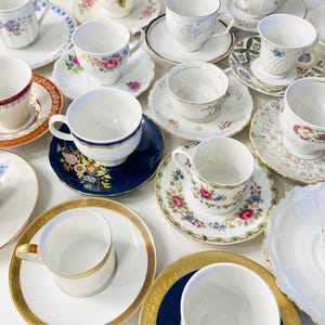 Puede incluir: Una colección de tazas de té y platillos vintage en varios diseños. Las tazas y los platillos presentan diseños florales, adornos dorados y una mezcla de colores, incluyendo blanco, azul, rosa y dorado. La disposición está sobre una superficie blanca.