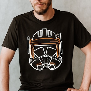 May include: A black t-shirt with a white outline of a Star Wars clone trooper helmet. The helmet has orange accents.