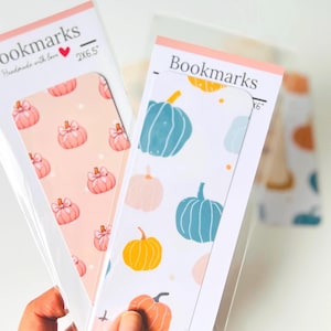 May include: Two bookmarks with pumpkin designs. One has a pink background with pink pumpkins, the other has a white background with blue, orange, and yellow pumpkins. The bookmarks are packaged in clear plastic.