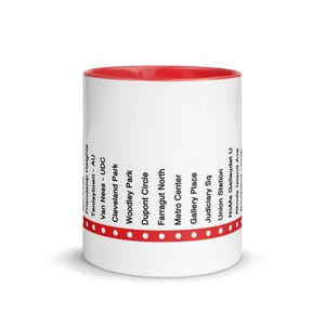May include: A white ceramic mug with a red interior and rim. The exterior features a horizontal red line with white dots, intersected by vertical black text listing Washington D.C. metro stations including Friendship Heights, Tenleytown, and Union Station.