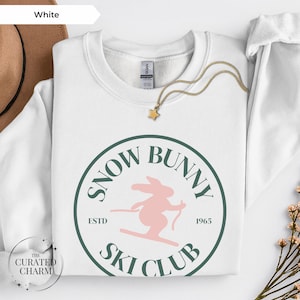 May include: White sweatshirt with a round, green and pink graphic that reads "Snow Bunny Ski Club ESTD 1965". A gold necklace with a star pendant rests on the sweatshirt. The sweatshirt is part of a clothing collection.