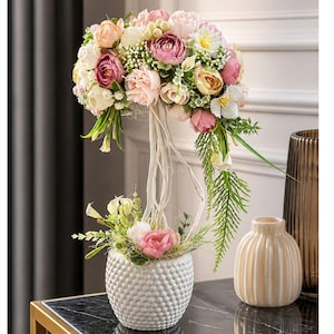May include: A decorative floral arrangement with artificial flowers in pink, white, and yellow. The arrangement is in a white textured vase, connected to a larger bouquet above. A small beige vase and a brown vase are also present.