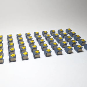 Op de afbeelding: Een verzameling kleine, rechthoekige spelstukken op een witte ondergrond. Elk stuk is een miniatuurboek met een blauwe kaft, beige pagina's en een geel embleem. De gelijkmatige opstelling suggereert een bordspelcomponent.