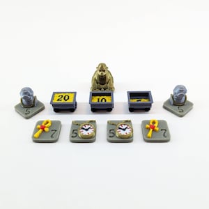 Puede incluir: Un conjunto de piezas de juego para un juego de mesa. Las piezas incluyen una figura de mono de oro, tres carros grises con monedas de oro, dos figuras grises con el número 5, y cuatro cuadrados grises con el número 5, un reloj, un ankh y el número 7.