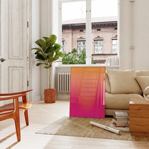 May include: A modern living room scene featuring a large, rectangular artwork with a vibrant gradient of orange to pink hues. The artwork leans against a wall near a window, complemented by a beige sofa, wooden chair, and a potted plant.