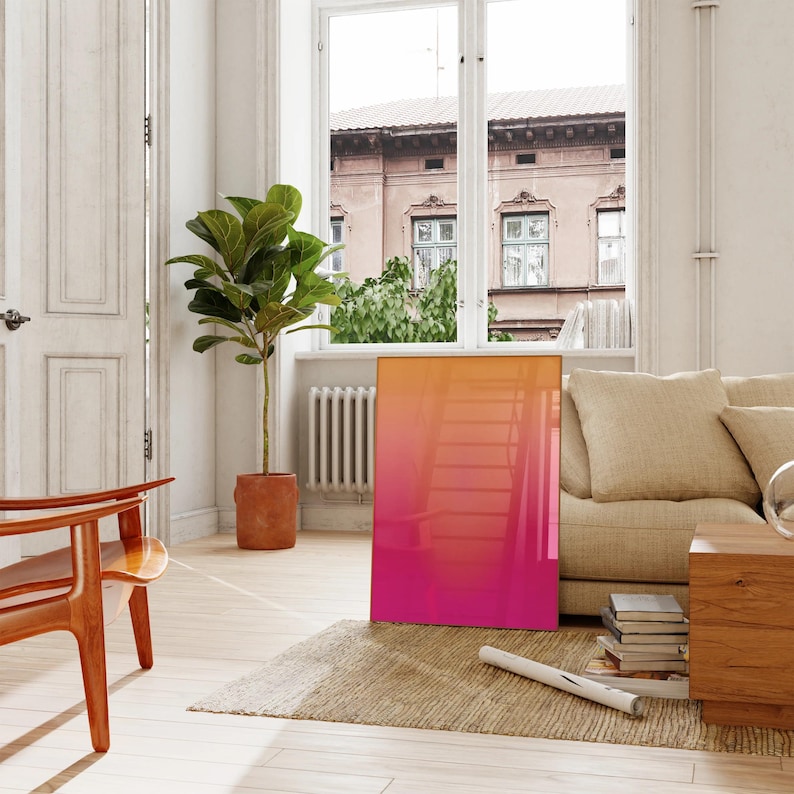 May include: A modern living room scene featuring a large, rectangular artwork with a vibrant gradient of orange to pink hues. The artwork leans against a wall near a window, complemented by a beige sofa, wooden chair, and a potted plant.