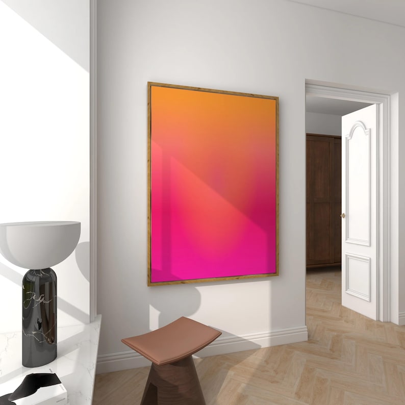 May include: A framed abstract art piece with a gradient of orange, pink, and red hues. The artwork is displayed on a white wall in a room with a wooden floor. A brown stool and a marble lamp are in the foreground.