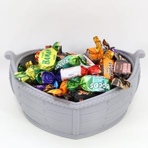 Puede incluir: Un cuenco gris en forma de barco lleno de caramelos de colores envueltos individualmente. El cuenco tiene un detalle de cuerda alrededor del borde y asas. Los caramelos son de varias marcas.