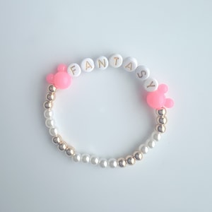 May include: A white and gold beaded bracelet with pink Mickey Mouse charms and the word "FANTASY" spelled out in gold letters on white beads.