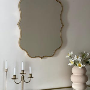 May include: A gold-toned, ornate mirror with a scalloped frame hangs on a white wall above a glass-top console table. A gold candelabra with four white candles sits on the table, along with a pink ceramic vase filled with white flowers.