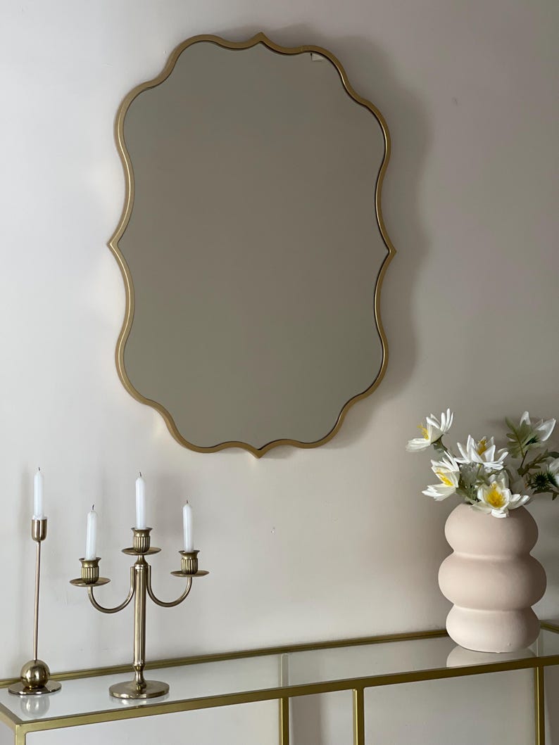 May include: A gold-toned, ornate mirror with a scalloped frame hangs on a white wall above a glass-top console table. A gold candelabra with four white candles sits on the table, along with a pink ceramic vase filled with white flowers.