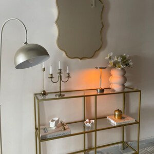 May include: A gold metal console table with glass shelves. The table has two shelves and is decorated with books, a vase of white flowers, and decorative objects. The table is in front of a white wall with a gold mirror.