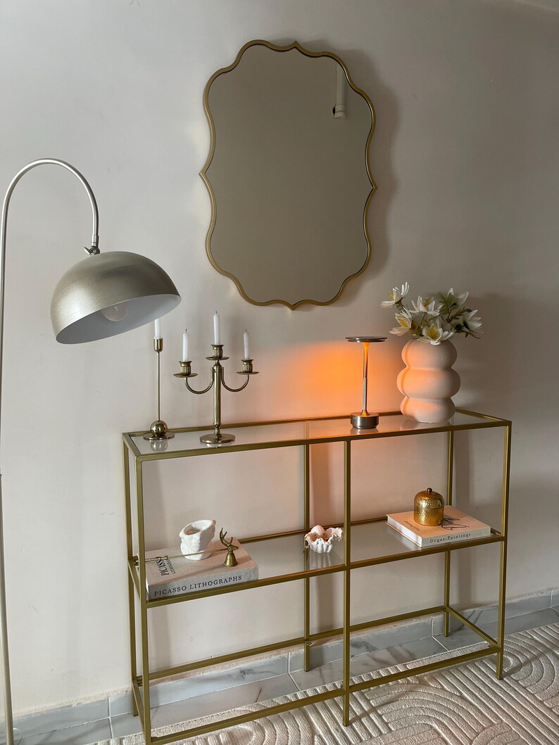 May include: A gold metal console table with glass shelves. The table has two shelves and is decorated with books, a vase of white flowers, and decorative objects. The table is in front of a white wall with a gold mirror.