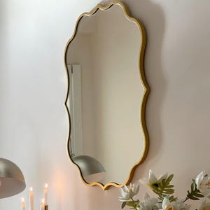 May include: A gold-framed, scalloped-edge mirror hangs on a white wall. The mirror reflects a white ceiling and a silver lamp.