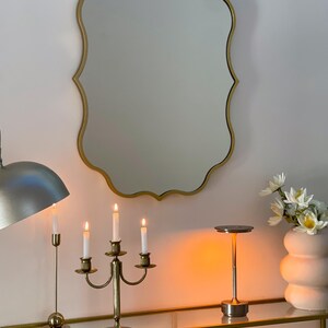 May include: A gold-toned, ornate, scalloped mirror hangs above a console table. The table has a glass top and a gold-toned base. A three-armed candelabra with lit candles sits on the table, along with a small, gold-toned lamp with a round shade.