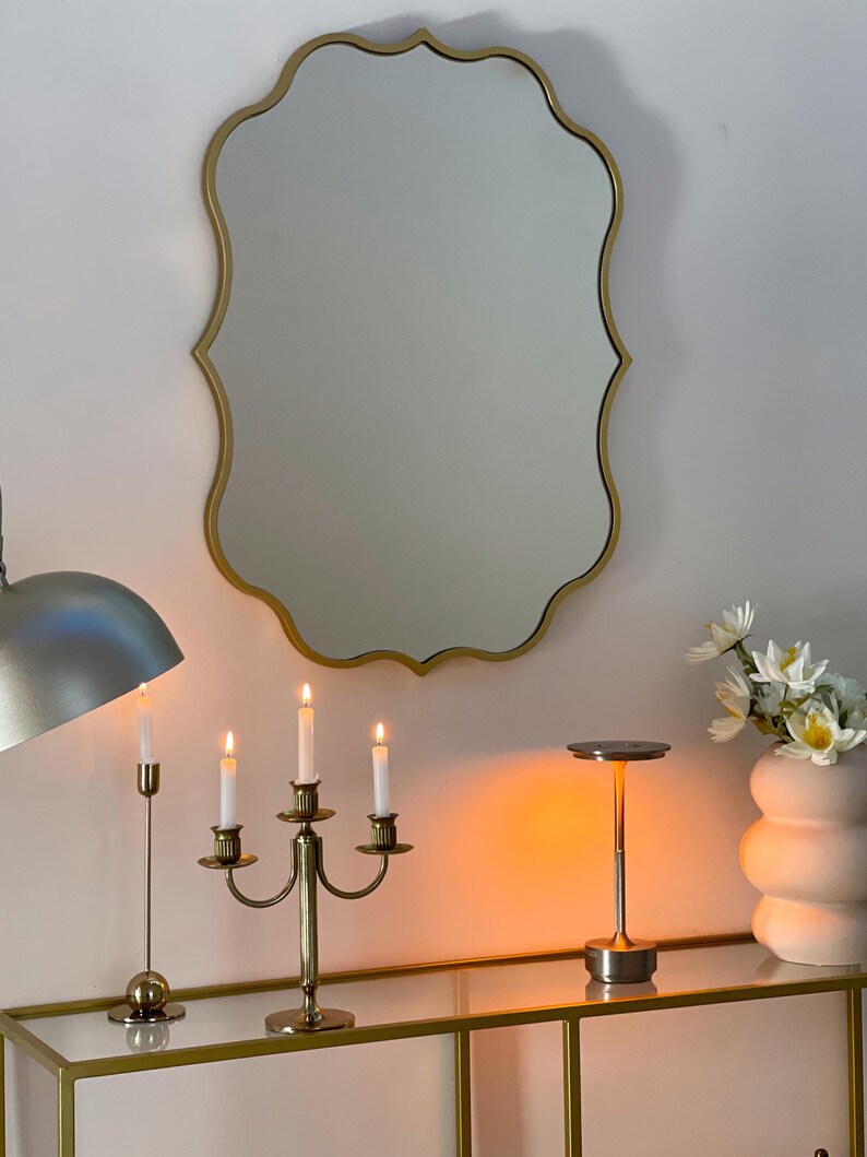 May include: A gold-toned, ornate, scalloped mirror hangs above a console table. The table has a glass top and a gold-toned base. A three-armed candelabra with lit candles sits on the table, along with a small, gold-toned lamp with a round shade.