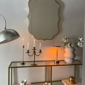 May include: A gold-framed mirror with a scalloped edge hangs above a glass shelf unit with gold metal framing. The shelf unit has two shelves and is decorated with candles, a vase of white flowers, and books. The books are titled "Picasso Lithographs" and "Degas Paintings".
