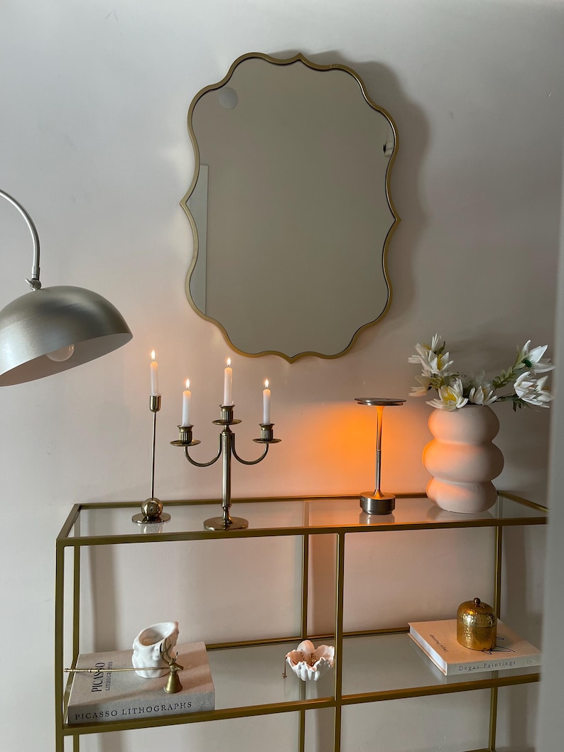 May include: A gold-framed mirror with a scalloped edge hangs above a glass shelf unit with gold metal framing. The shelf unit has two shelves and is decorated with candles, a vase of white flowers, and books. The books are titled "Picasso Lithographs" and "Degas Paintings".