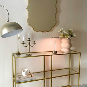 May include: A gold metal console table with glass shelves. The table has a white ceramic vase with white flowers, a gold candlestick holder, and a gold table lamp with a white shade. The table is sitting on a white rug with a geometric pattern.