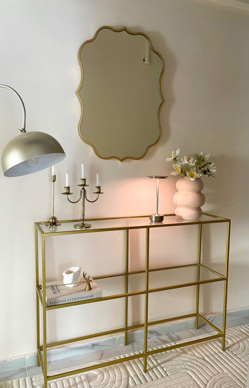 May include: A gold metal console table with glass shelves. The table has a white ceramic vase with white flowers, a gold candlestick holder, and a gold table lamp with a white shade. The table is sitting on a white rug with a geometric pattern.
