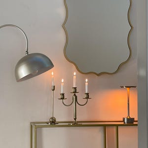 May include: A gold-framed, scalloped mirror hangs above a gold metal shelf with a gold candlestick holder with four lit candles, a silver lamp, and a book titled "Picasso Lithographs".