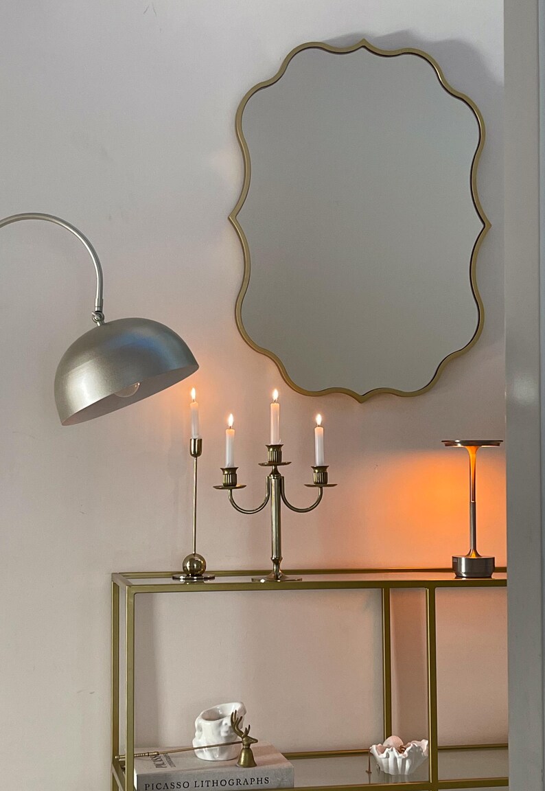 May include: A gold-framed, scalloped mirror hangs above a gold metal shelf with a gold candlestick holder with four lit candles, a silver lamp, and a book titled "Picasso Lithographs".