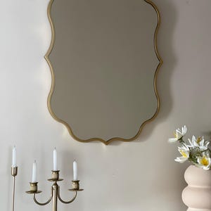 May include: A gold-framed, scalloped mirror hangs on a white wall above a glass shelf with a gold metal candlestick and a pink ceramic vase with white flowers.