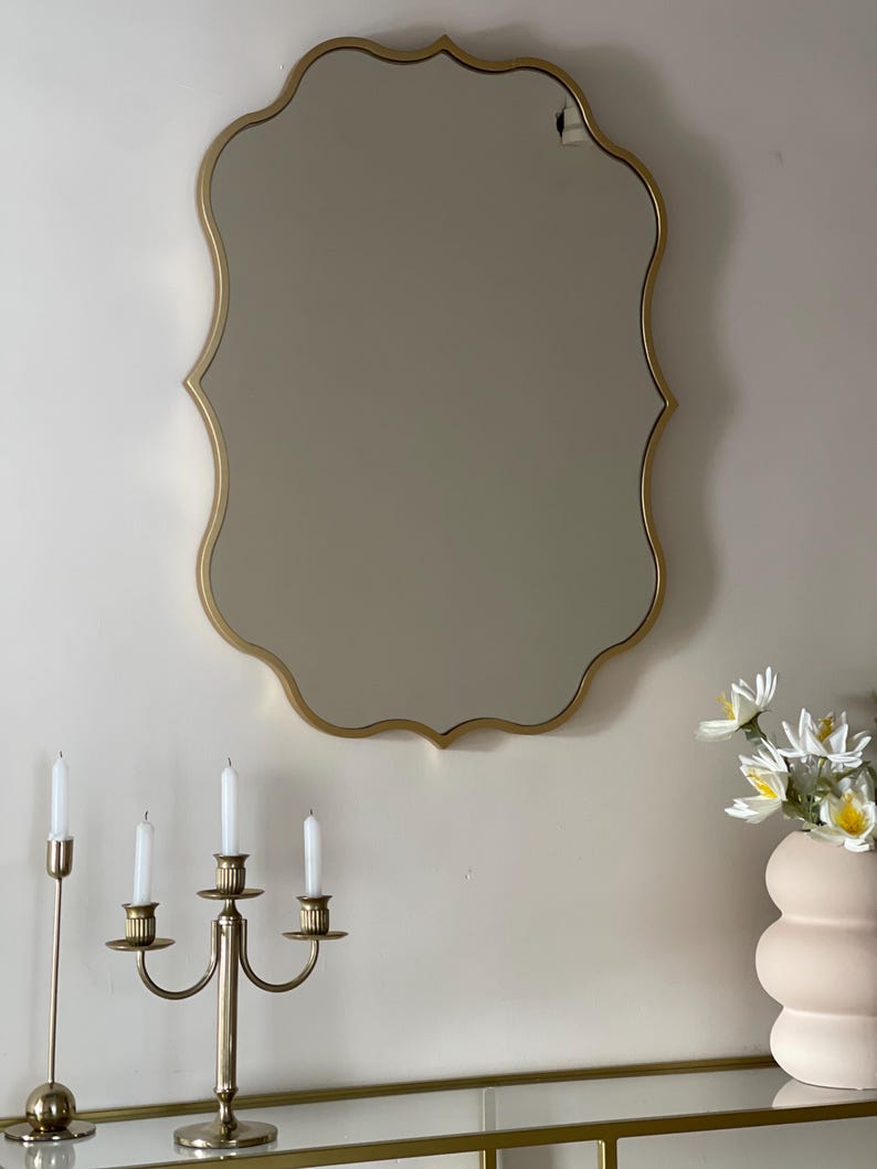 May include: A gold-framed, scalloped mirror hangs on a white wall above a glass shelf with a gold metal candlestick and a pink ceramic vase with white flowers.