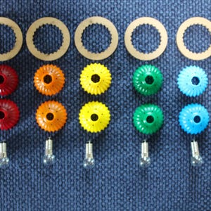 May include: A set of five light fixture components arranged in rows. Each column features a tan circular ring, two fluted plastic shades in red, orange, yellow, green, or blue, and a small clear glass light bulb with a silver metal base.