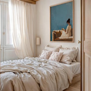 May include: A bedroom scene featuring a bed with beige bedding and decorative pillows. A framed painting of a person reading sits above the bed. Sheer curtains frame a window, and a wall-mounted lamp is visible.