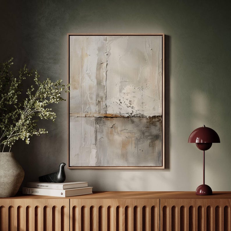May include: Abstract art piece in a wooden frame, featuring a textured, neutral color palette of white, beige, and gray. A burgundy table lamp and decorative items sit on a wooden cabinet. A vase with greenery is on the left.