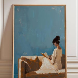 May include: A framed art print depicts a person reading on a sofa. The artwork features a person in a white dress, seated on a brown sofa, against a blue background. The print is encased in a wooden frame.
