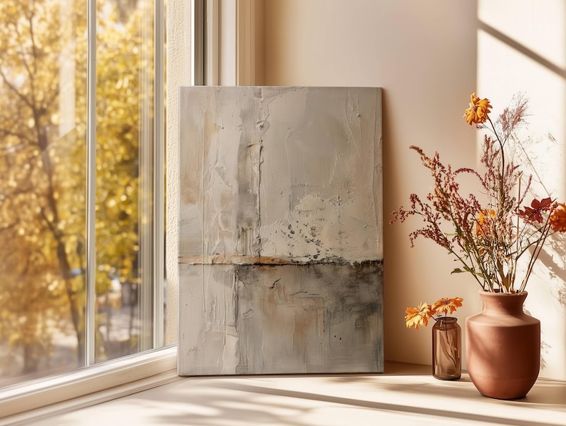 May include: Abstract art piece with a textured, neutral color palette. The artwork is displayed near a window with a view of fall foliage. A terracotta vase and a small glass bottle hold dried flowers, adding a touch of warmth to the scene.