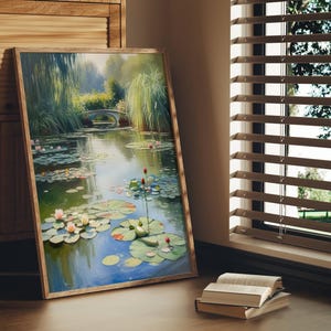 Puede incluir: Una pintura enmarcada de una serena escena de estanque con nen&uacute;fares, un peque&ntilde;o puente y exuberante vegetaci&oacute;n. La obra de arte se exhibe sobre un suelo de madera, junto a un mueble de madera y una ventana con persianas. Una pila de libros descansa en el suelo.
