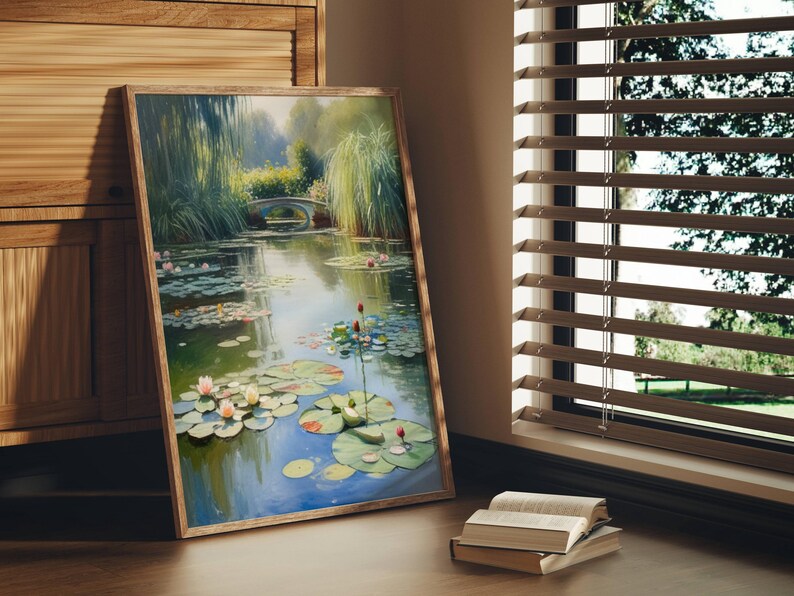 Puede incluir: Una pintura enmarcada de una serena escena de estanque con nen&uacute;fares, un peque&ntilde;o puente y exuberante vegetaci&oacute;n. La obra de arte se exhibe sobre un suelo de madera, junto a un mueble de madera y una ventana con persianas. Una pila de libros descansa en el suelo.