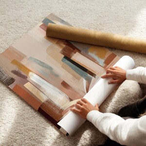 May include: An abstract art print with brushstroke designs in shades of brown, beige, and blue. A rolled-up print and a cardboard tube are next to the artwork. The print is on a light-colored carpet.