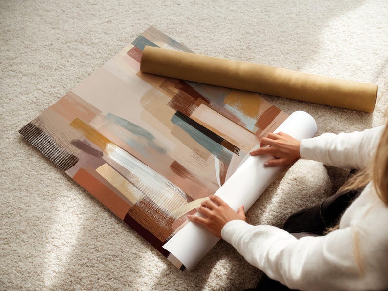 May include: An abstract art print with brushstroke designs in shades of brown, beige, and blue. A rolled-up print and a cardboard tube are next to the artwork. The print is on a light-colored carpet.