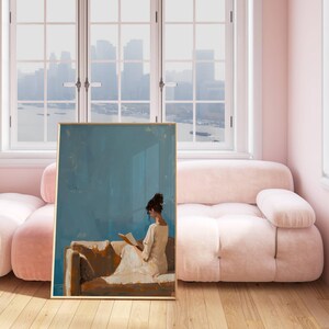 May include: A framed art print depicts a person reading, set against a blue background. The artwork is displayed in a room with a pink sofa and walls, and a large window overlooking a cityscape. The print has a gold frame.