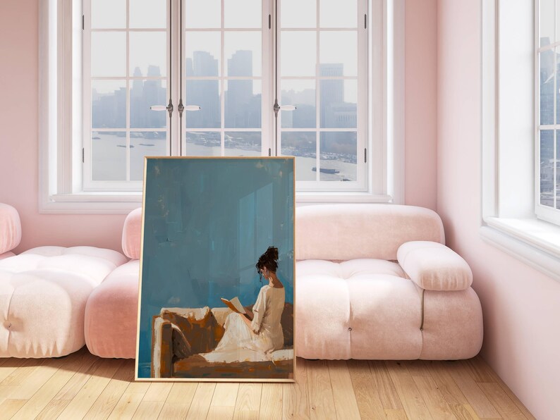 May include: A framed art print depicts a person reading, set against a blue background. The artwork is displayed in a room with a pink sofa and walls, and a large window overlooking a cityscape. The print has a gold frame.