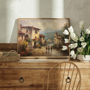 Pu&ograve; includere: Un dipinto incorniciato di un pittoresco villaggio italiano, su una credenza in legno rustico con un cesto di vimini e un vaso di tulipes bianchi.