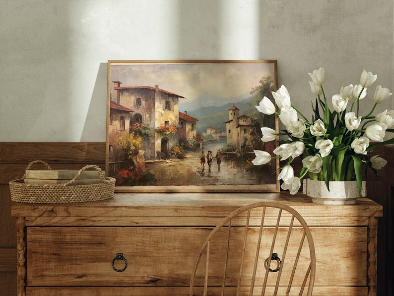 Pu&ograve; includere: Un dipinto incorniciato di un pittoresco villaggio italiano, su una credenza in legno rustico con un cesto di vimini e un vaso di tulipes bianchi.