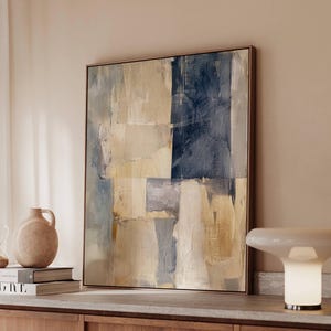 May include: Abstract art print in a wooden frame. The artwork features a textured, layered design with blocks of beige, navy blue, and gray. The painting is displayed on a wooden cabinet, with a vase and books visible.