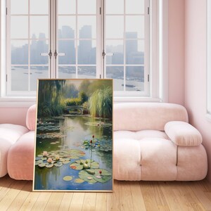 Puede incluir: Una pintura enmarcada representa una serena escena de estanque con nen&uacute;fares, un peque&ntilde;o puente y exuberante vegetaci&oacute;n. La obra de arte se exhibe en una habitaci&oacute;n con un sof&aacute; rosa y una gran ventana con vistas a un paisaje urbano. La pintura tiene un marco dorado.
