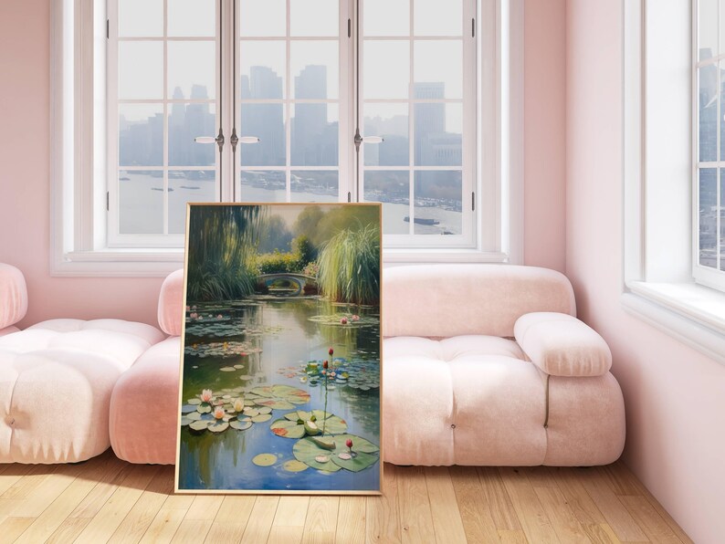 Puede incluir: Una pintura enmarcada representa una serena escena de estanque con nen&uacute;fares, un peque&ntilde;o puente y exuberante vegetaci&oacute;n. La obra de arte se exhibe en una habitaci&oacute;n con un sof&aacute; rosa y una gran ventana con vistas a un paisaje urbano. La pintura tiene un marco dorado.