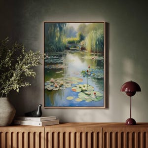 Puede incluir: Una pintura enmarcada representa una serena escena de estanque con nen&uacute;fares, un peque&ntilde;o puente y exuberante vegetaci&oacute;n. La obra de arte se exhibe sobre un mueble de madera, complementada con un jarr&oacute;n con ramas y una l&aacute;mpara de mesa burdeos.