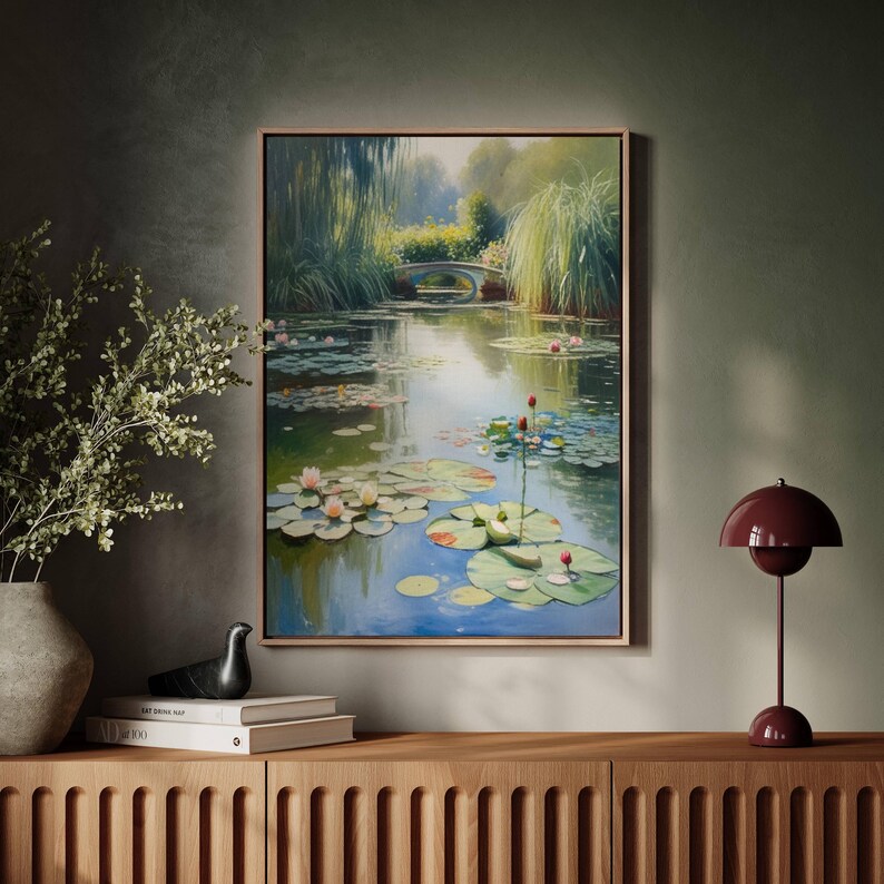 Puede incluir: Una pintura enmarcada representa una serena escena de estanque con nen&uacute;fares, un peque&ntilde;o puente y exuberante vegetaci&oacute;n. La obra de arte se exhibe sobre un mueble de madera, complementada con un jarr&oacute;n con ramas y una l&aacute;mpara de mesa burdeos.